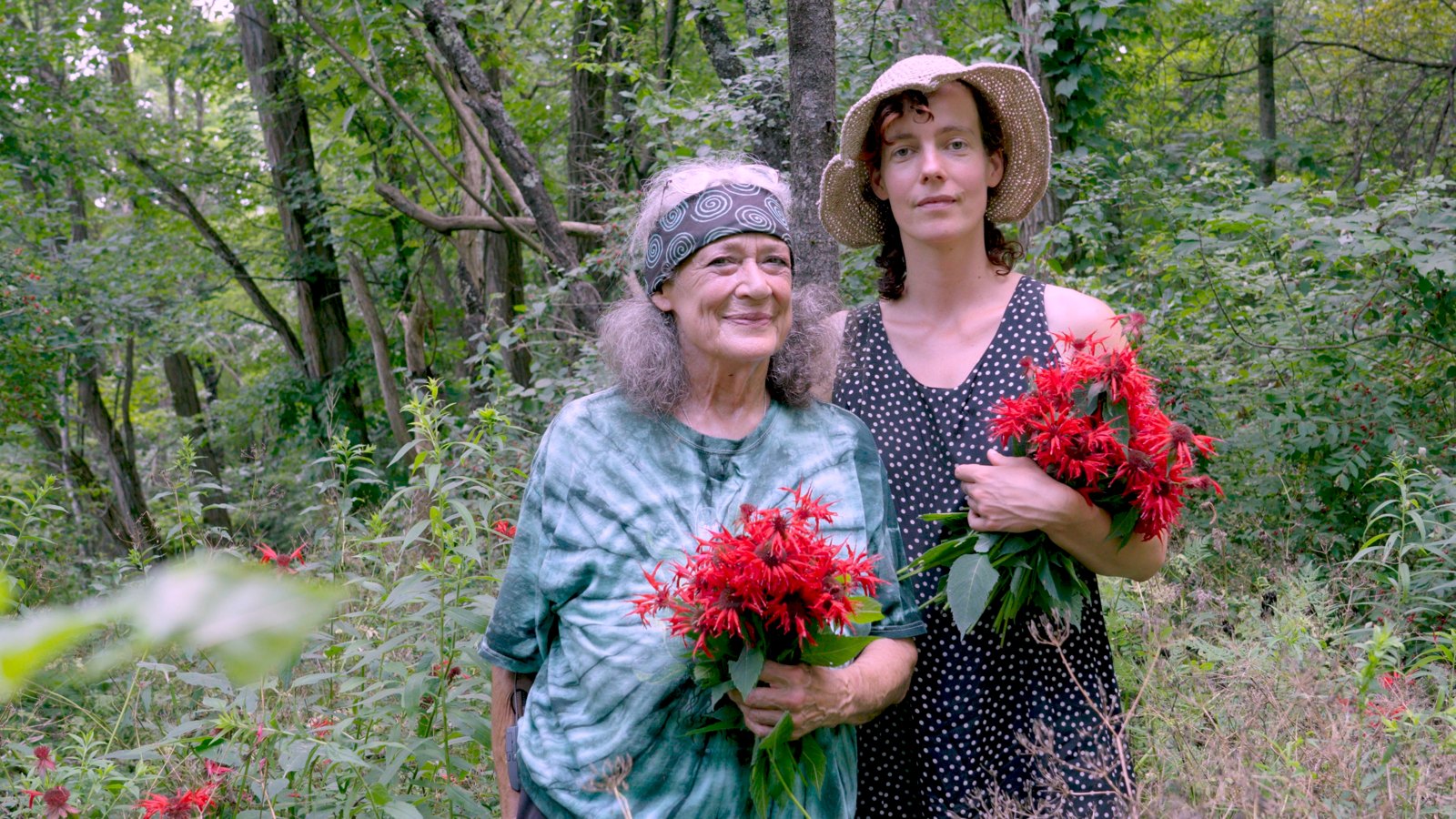 Still from the film: Susun Weed and her apprentice Julie standing together in a sunlit forest clearing, each holding a bouquet of bright red bee balm.